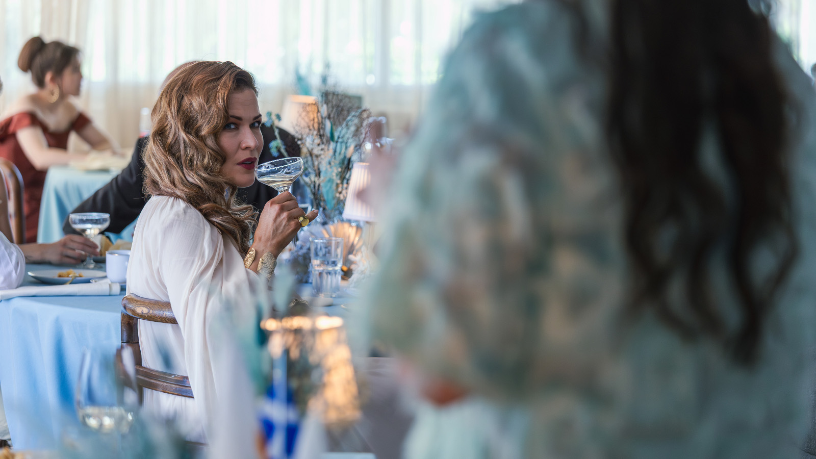 Kuvassa on ravintolatila ja oikealla edessä on himmeästi seisomassa nainen, josta näkyy turkoosin valkoista puseroa ja hieman pitkiä hiuksia selässä. Vasemmalla kauempana istuu Jenni Vartiainen sivuttain ja hän katsoo tulijaa sekä siemaileen oikeassa kädessään olevaa drinkkiä.