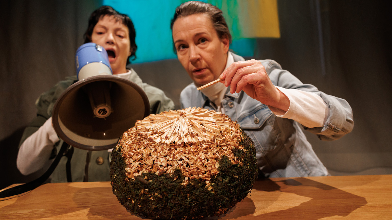 Kuvassa ovat istumassa pöydän ääressä Teija Töyry kädessään megafoni ja Heidi Syrjäkari ojentamassa vasenta kättään kohti pöydällä olevaa isoa palloa. Se on ruskea ja siinä on kultausta päällä. 