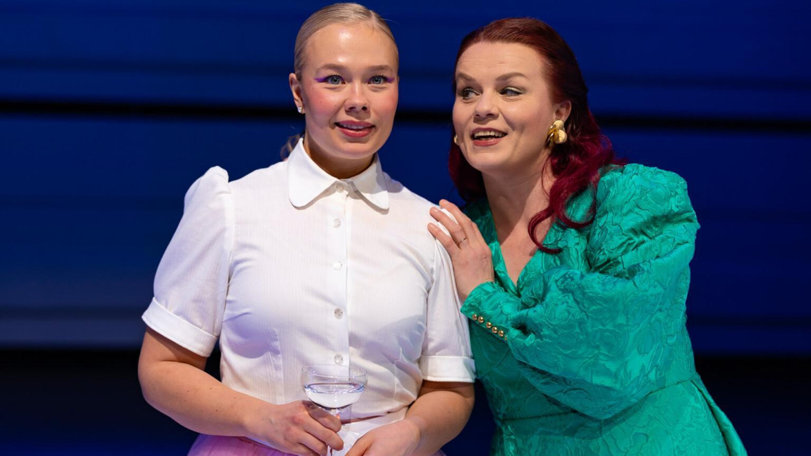 Kuvassa on Ronja Keiramo valkoisessa puserossa sampanja lasi kädessään ja oikealla on Ella Mustajärvi vihreässä mekossa. He ovat seisomassa puolivartalokuvassa. Tausta on tumman sininen.