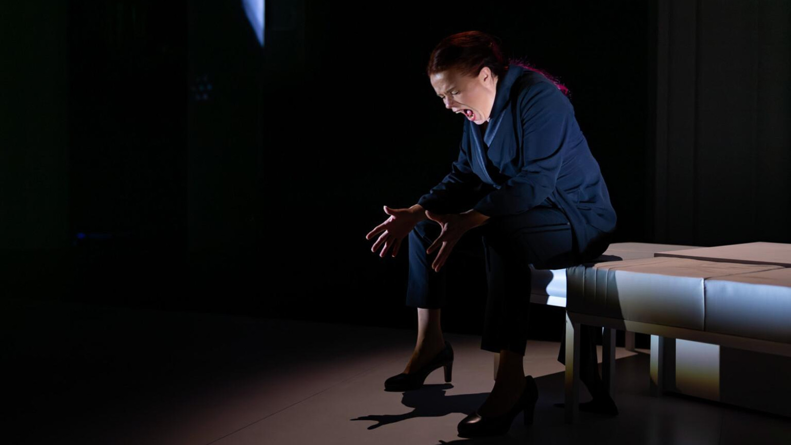 Kuvassa on Ella Mustajärvi istumassa tuskaisen näköisenä säntyn laidalla. Hänellä on tumman sininen mekko päällään. Tausta on tumma.