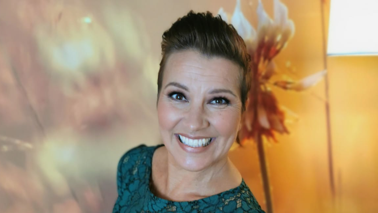 Kuvassa on Arja Koriseva puolivartalokuvassa ja hänellä on hiukset takana ja niissä valkoinen kukka puolittain takana. Hänellä on siniharmaa mekko päällään. Taustalla on vaalean punertavan vaalea teos ja oikealla oranssia.