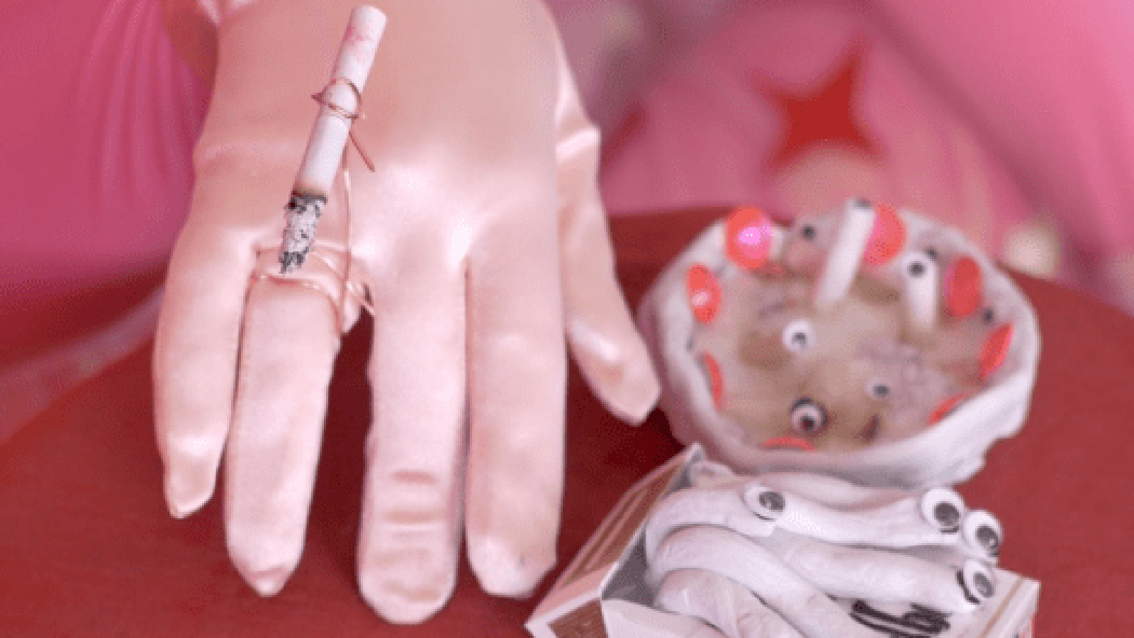 Kuvassa on pinkillä silkkisellä pohjalla käsi vaaleassa hansikkaassa ja sen päälle on kiinnitetty tupakka. Oikealla on epämääräisen muotoinen valkoinen tuhkakuppi, jossa on vaaleita keraamisia tumppeja. Edessä käyristettyjä keraamisia tupakoita.