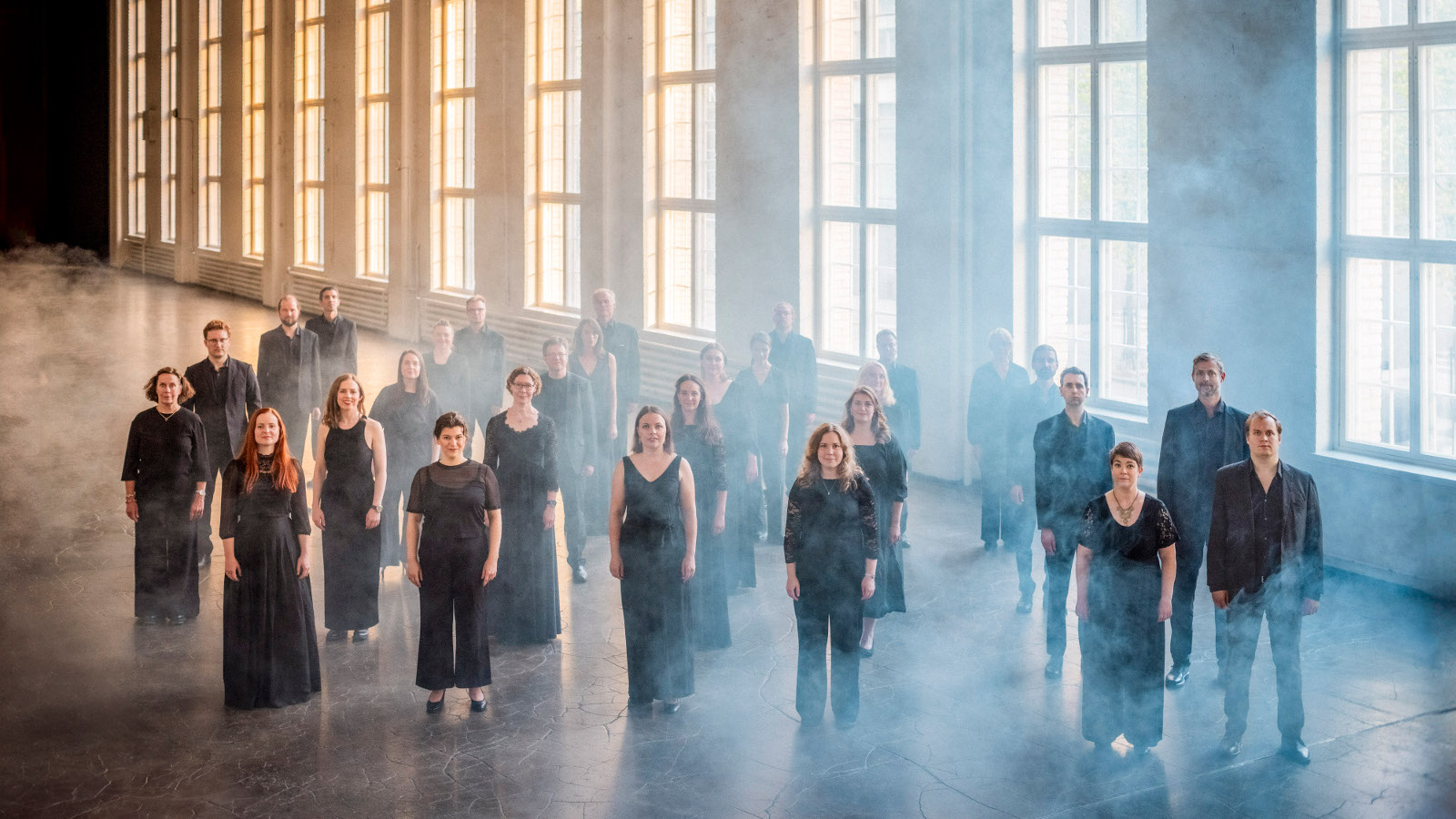 Kuvassa on Helsingin kamarikuoron jäsenet seisomassa mustissa mekoissa ja puvuissa. He ovat isossa hallissa, jossa on korkeat ruutuikkunat rivissä, joista tulee valoa.  