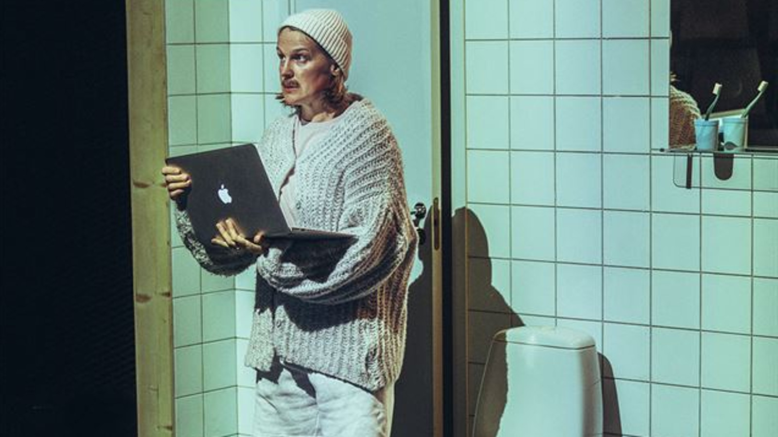 Kuvassa on nuori miesvaaleassa villapaidassa seisomassa Wc-tilassa ja hänellä on läppäri kädessään. Wc:n kaakelit ovat valkoisia ja ovi sekä wc-istuin.