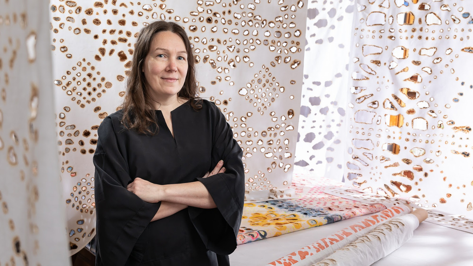 Kuvassa on vasemmalla Johanna Pöykkö mustassa mekossa puolivartalokuvassa. Taustalla on ripustettu kankaita, joissa on ruskeareunaisia reikiä erilaisina kuvioina.