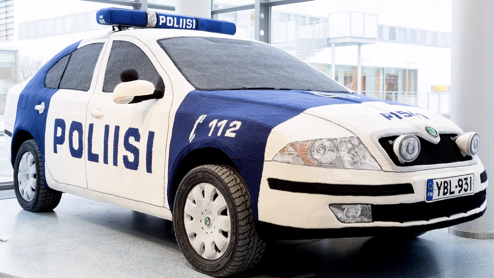 Kuvassa on sisätilassa poliisiauto vinottain. Sen väri on valkoinen ja sininen.  Vasemmassa kyljessä lukee suurella Poliisi ja sinisellä pohjalla numero 112. Kuvassa on tekstiilityö.
