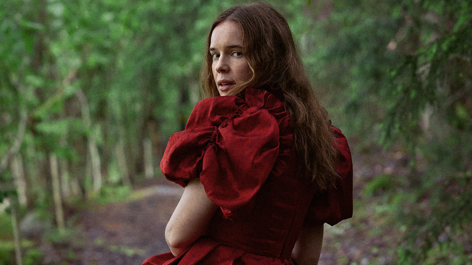 Kuvassa on Marketta Tikkanen selin kävelemässä metsätietä, jonka varrella on koivuja. Tkkasella on punainen mekko päällään ja siinä on puhvihihat.