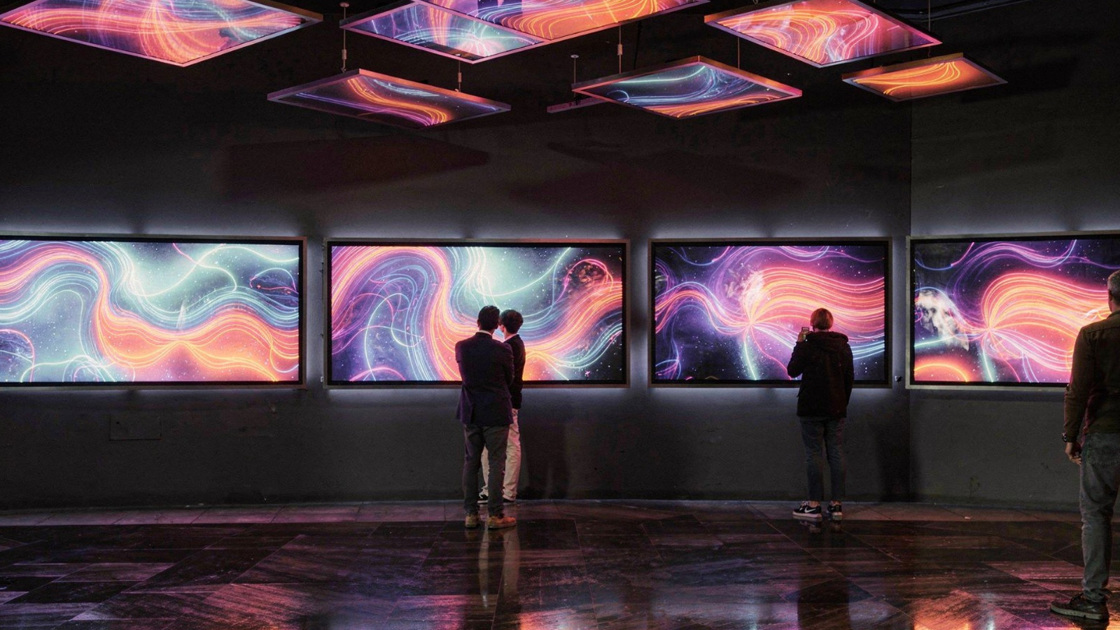 Kuvassa on keskellä ruuturivissä värikkäitä kiemurtelevia viivoja ja pintoja. Tila on tumma ja kuvat heijastuvat kattoon ja lattiaan. Kuvia katselee pariskunta ja yksittäinen katselija.