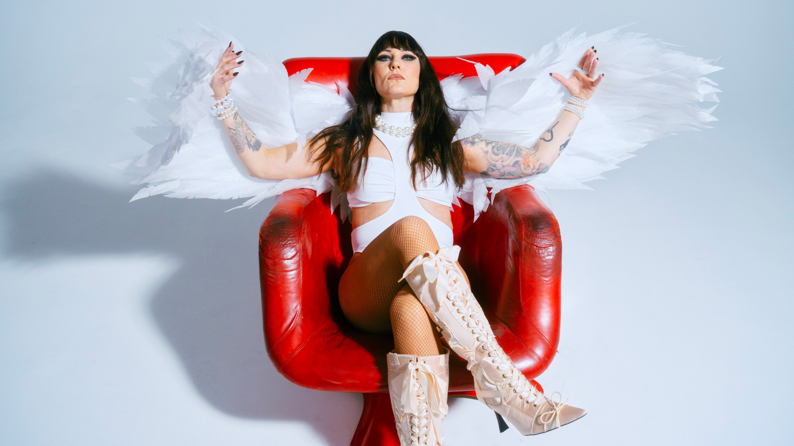 Kuvassa on Mira Luoti istumassa punaisessa kiiltävässä nojatuolissa. Hänellä on valkoinen mekko ja kädet levitettyinä sivuille ja niissä on enkelin siivet. Tausta on vaalea ja siinä on varjo vasemmalla.