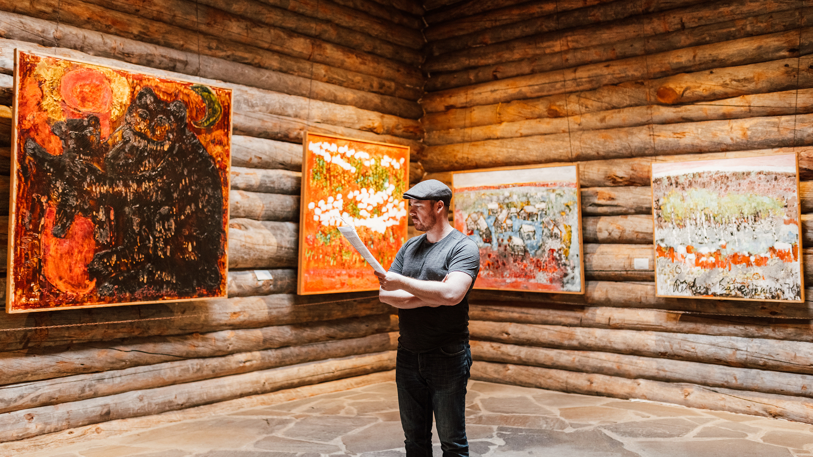 Kuvassa on ruskealla hirsiseinällä neljä oranssisävyistä teosta nurkittain. Nurkan kohdalla seisoo mies katsomassa teoksia. 