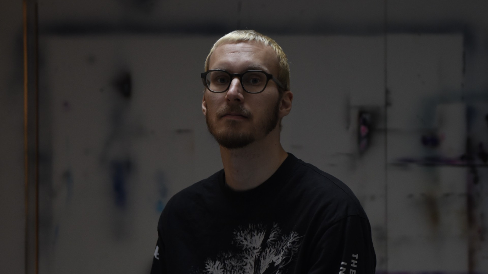 Kuvassa on Roope Itäranta puolivartalokuvassa. Hänellä on musta t-paita päällään. Hänellä on lyhyet vaaleat hiiukset. Kuvan sävy ja tausta on tumma.