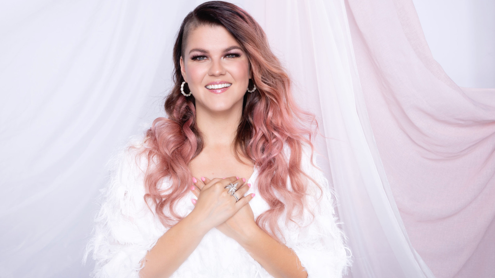 Kuvassa on Saara Aalto puolivartalokuvassa ja hänellä on valkoinen, pörröinen pusero päällään. Hänellä on punertavat kiharaiset hiukset ja hän pitää ksiään rinnallaan. Tausta on valkoinen.