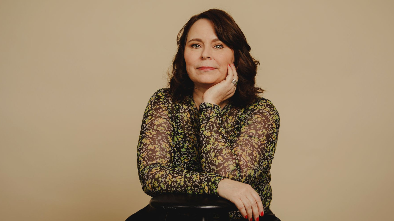 Kuvassa on Saila Susiluoto ja hän istuu sekä nojaa tuolin selkämykseen. Hänellä on ruskean mustan kirjava puseoro päällään.  Kuvan tausta on ruskehtavan harmaa. 