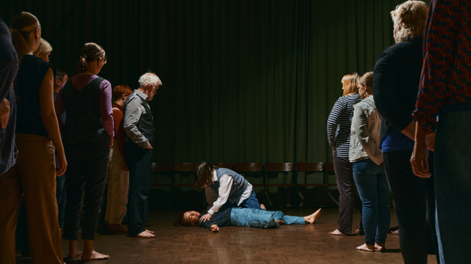 Kuvassa on keskellä lattialla makaava nainen ja häntä hoitaa toinen nainen. Molemmin puolin seisoo jonossa miehiä ja naisia ja heillä on tavalliset vaatteet. Tausta on tumma.