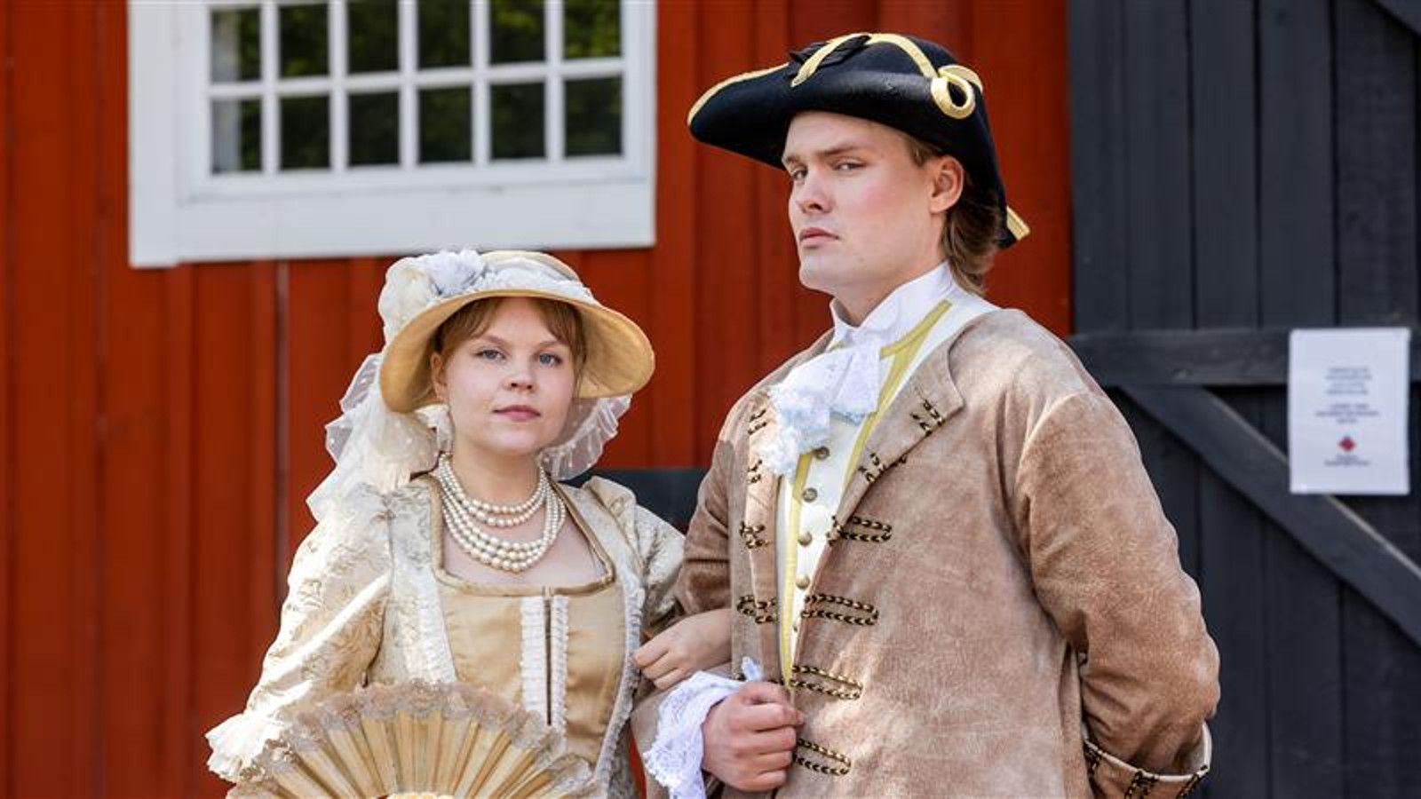 Kuvassa on vasemmalla nuori nainen vaalean ruskeassa vanhanaikaisessa mekossa ja lierihatussa. Asussa ja hatussa on valkoisia röyhelöitä ja brodyyrejä. Oikealla on nuori mies, jolla on musta aatelisen suikkahattu päässään sekä koristeellinen vaalean ruskea takki päällään ja valkoinen röyhelöpaita. Taustalla on punaisen puurakennuksen seinää ja valkoinen, pieniruutuinen ikkuna.