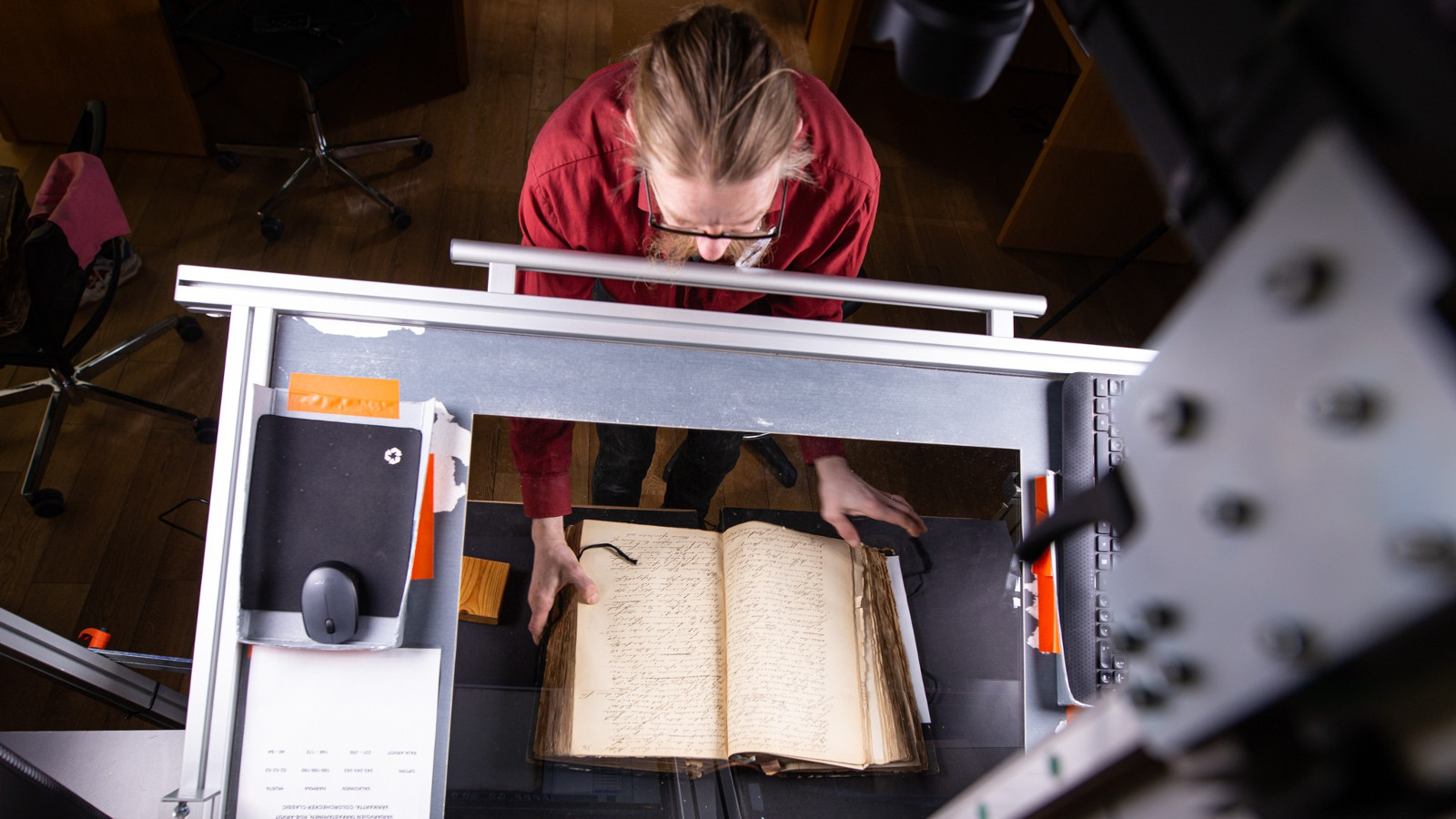 Kuvassa on ylhäältä päin kuvattuna punaisessa puserossa oleva mies, joka pitelee kirjaa alustalla. Päällä on harmaa metallikehikko, jolla digitoidaan kirjaa.