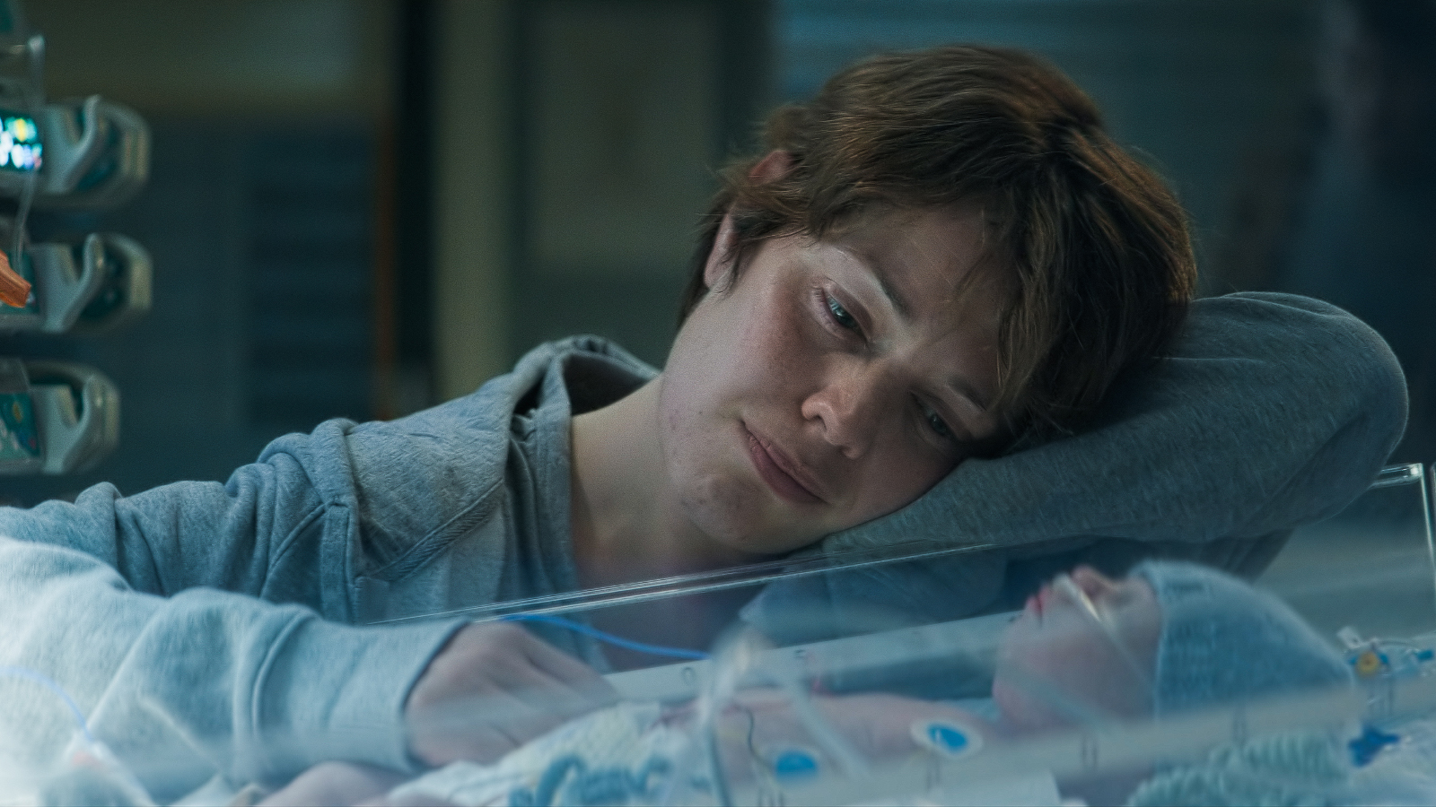 Kuvassa on Oona Airola, joka katselee keskoskaapissa nukkuvaa vauvaa. Hänellä on vaalean sininen pusero päällään.