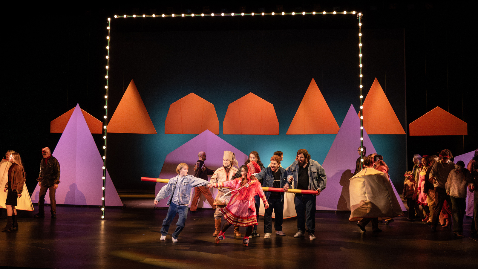 Kuvassa on tummalla näyttämöllä takana punaisia kartiomaisia telttoja. alemana on näytteljöitä tanssimassa edessä värikkäissä vaatteissa ja taaempana seisoo harmassa puvuissa olevia näyttelijöitä. Vasemmalla on kartiomainen lila teltta.