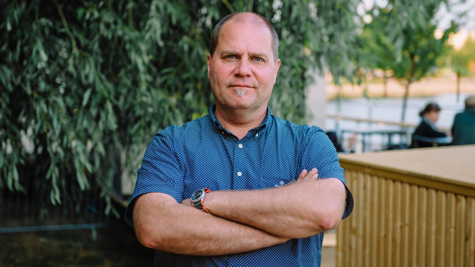 Kuvassa on keskellä Stuba Nikula seisomassa puolivartalokuvassa. Hänellä on sininen, lyhythihainen paita päällään ja hänellä on kädet toistensa päällä. Taustalla on alaspäin laskeutuvia lehtipuun oksia.