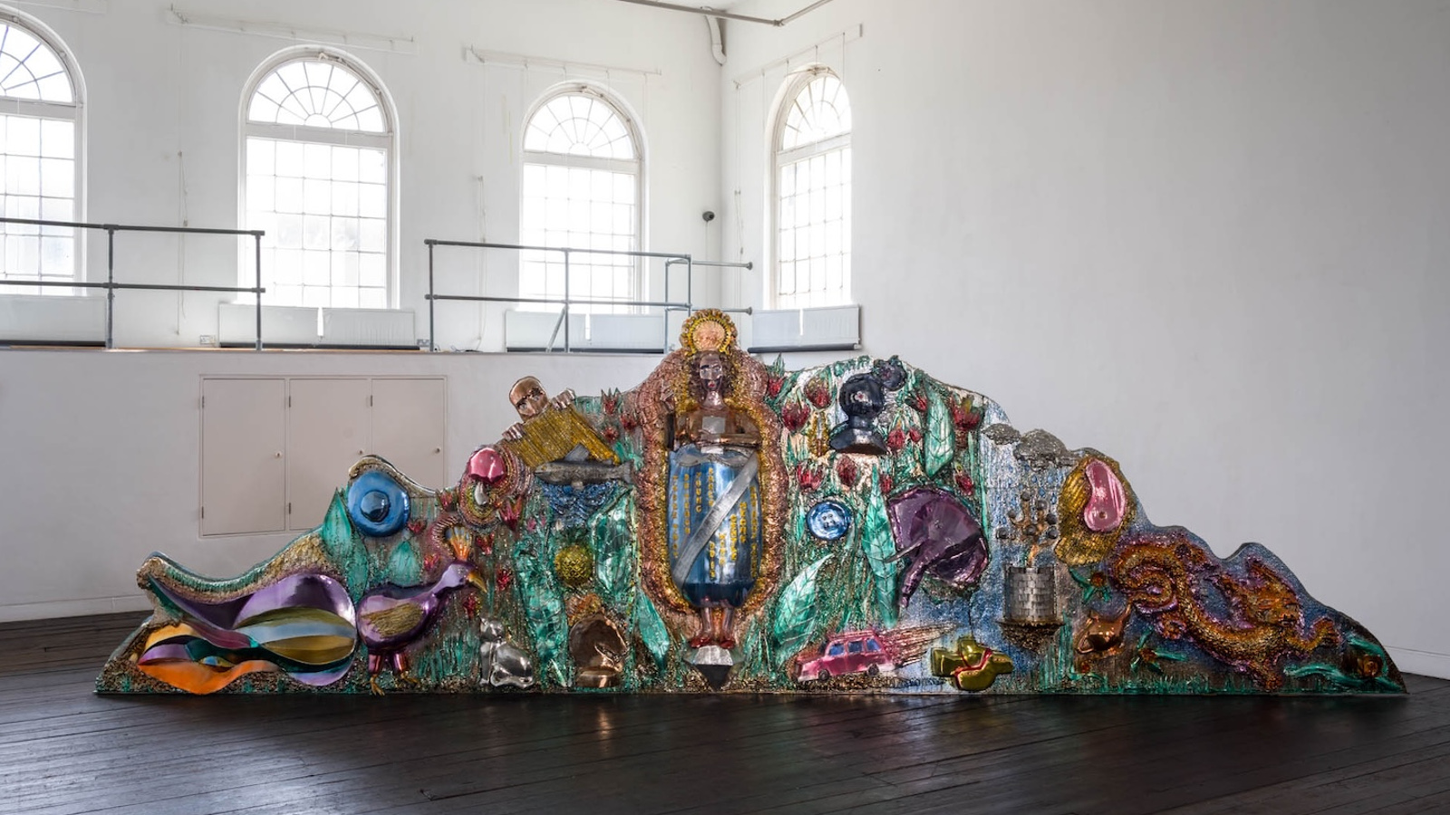 Kuvassa on isossa valkoisessa tilassa, jossa on vasemmalla ylhäällä isoja, yläosasta pyöreitä ja pikkuryytuisia ikkunoita. Tummalla lattialla on kiiltävävärisiä, erityisesti turkoosia, ruskeaa ja lilaa ison krokotiilimaisen hahmon päällä.  Hahmo nousee ylöspäin ja laskeutuu alas.  Päällä on keraamisia kuvioita ja mm. lintuja.