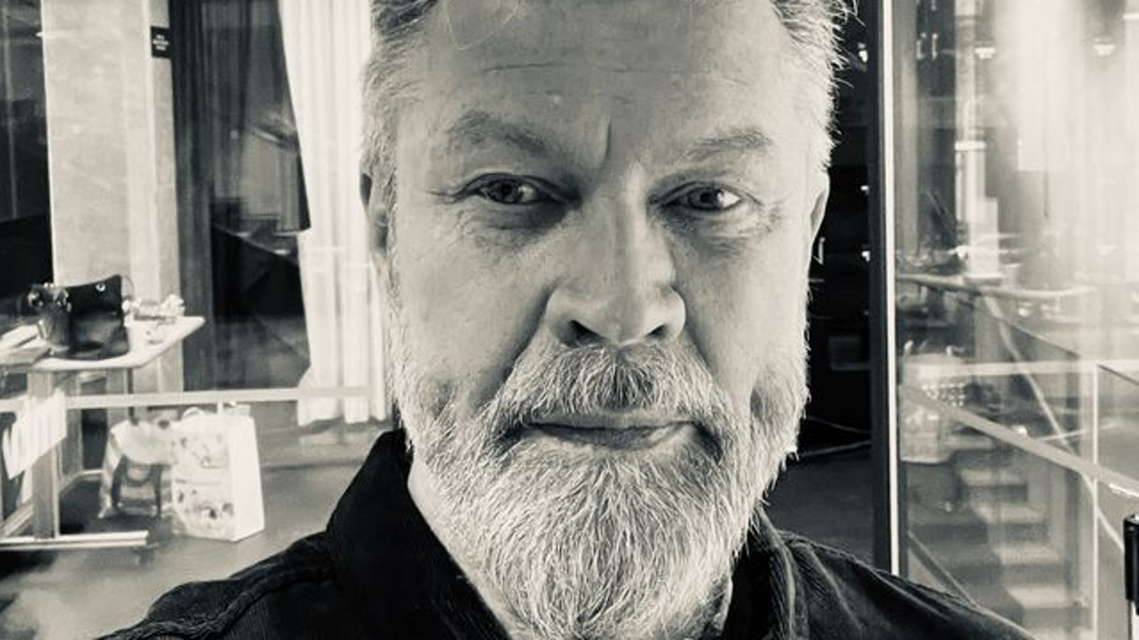 Kuvassa on Asko Kallosen kasvokuva ja hänellä vaalea poskiparta. Taustalla on huonekaluja. Kuva on musta-valkoinen. 