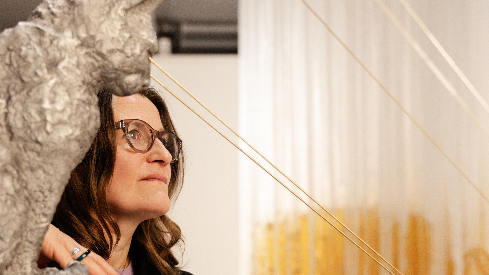 Kuvassa on vasemmalla on harmaa kissaveistoja sen takaa näkyy Hanna Vihriälän kasvot sivuttain. Hänellä on tummat hiukset ja silmälasit. Oikealla on valkoista verhoa, jossa on alhaalla keltaista. 