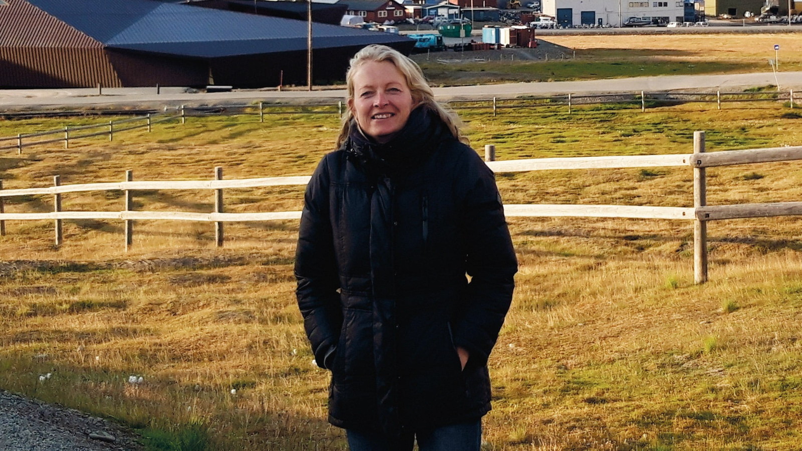 Kuvassa on keskellä Jessica Haapkylä seisomassa mustassa takissa. Taustalla on vaalean keltaista peltoa, jossa on puuaitausta ja takana kaksi isoa telttaa. 