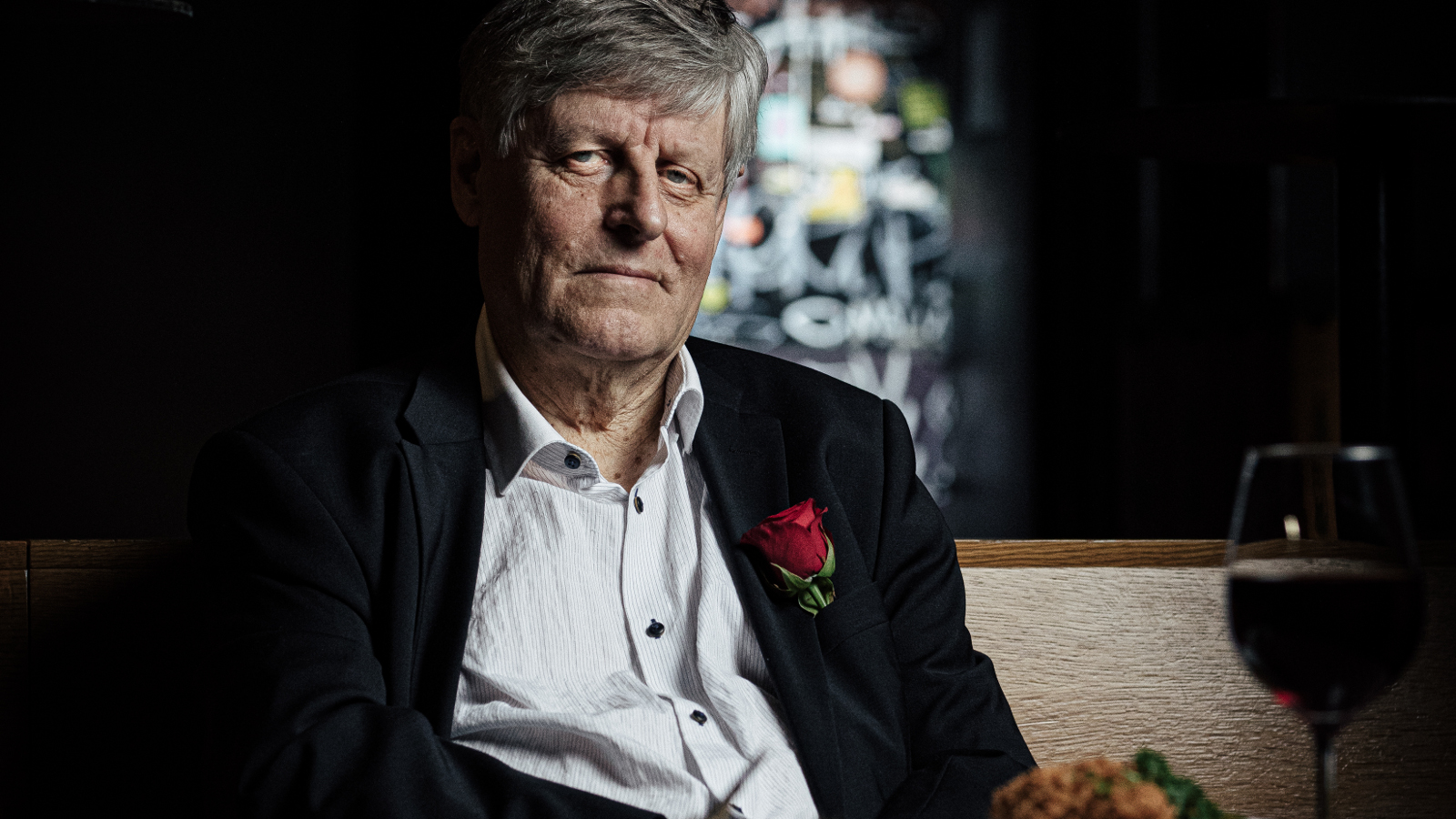 Kuvassa on Juhani Merimaa puolivartalokuvassa ja hän istuu. Hänellä on musta takki ja valkoinen paita. Tausta on melko tumma sekä vasemmalla että oikealla.