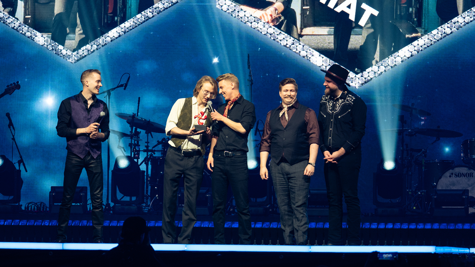  Kuvassa ovat lavalla Komiat-yhtyeen viisi miesjäsentä.  Heillä on mustat esiintymisaset ja yhdellä on valkoinen paita ja musta liivi. Taustalla on sinistä valaistusta.