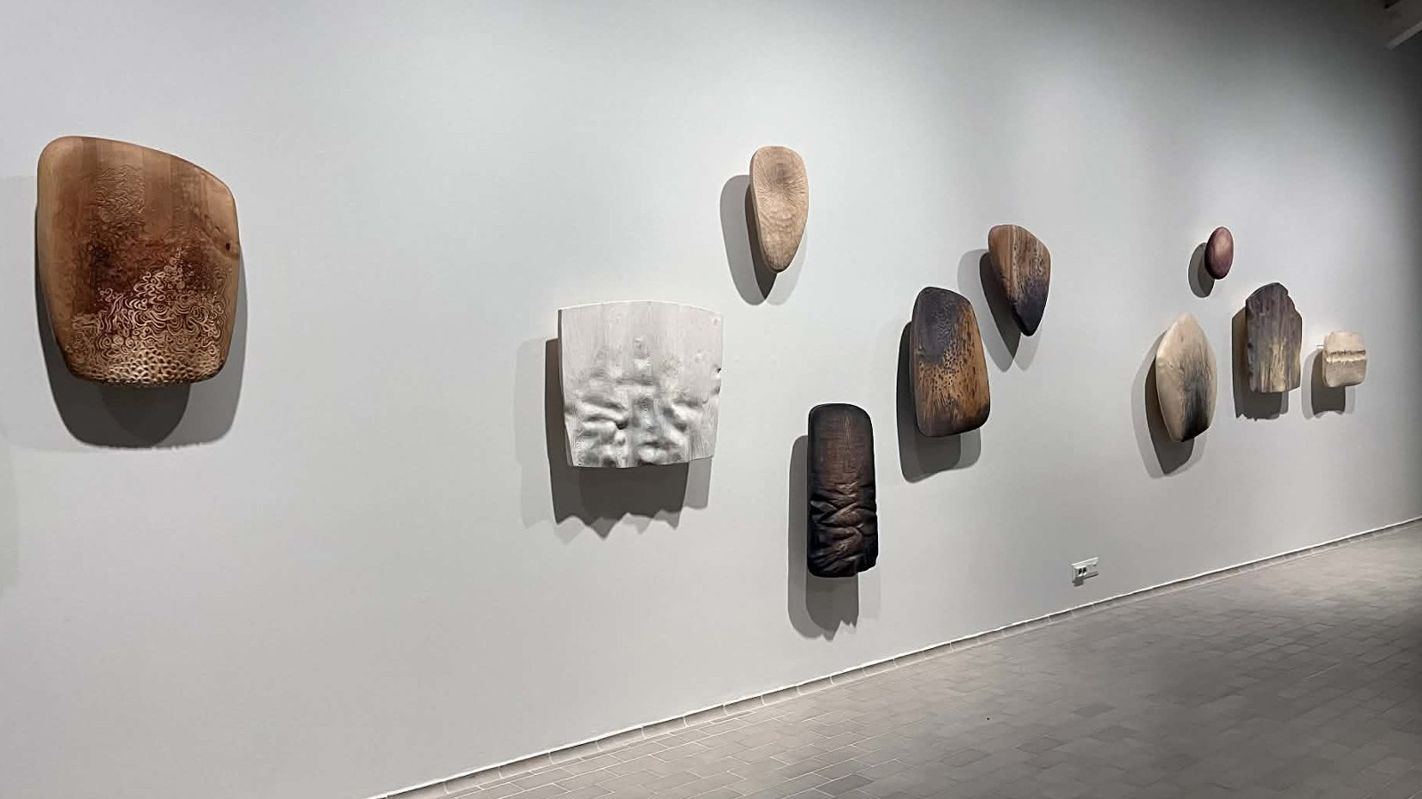 Kuvassa on vaalean harmaalla seinällä, jossa on ruskehtavia ja vaaleita eri värisiä ja muotoisia, melko pieniä seinäreliefejä.