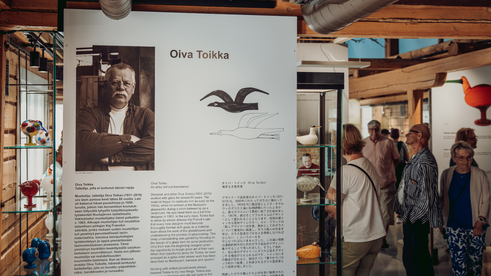 Kuvassa on vasemmalla edessä iso infotaulu, jossa on Oiva Toikan kuva ja alla tekstiä. Oikealla taaempana on ihmisiä katsomassa näyttelyä.