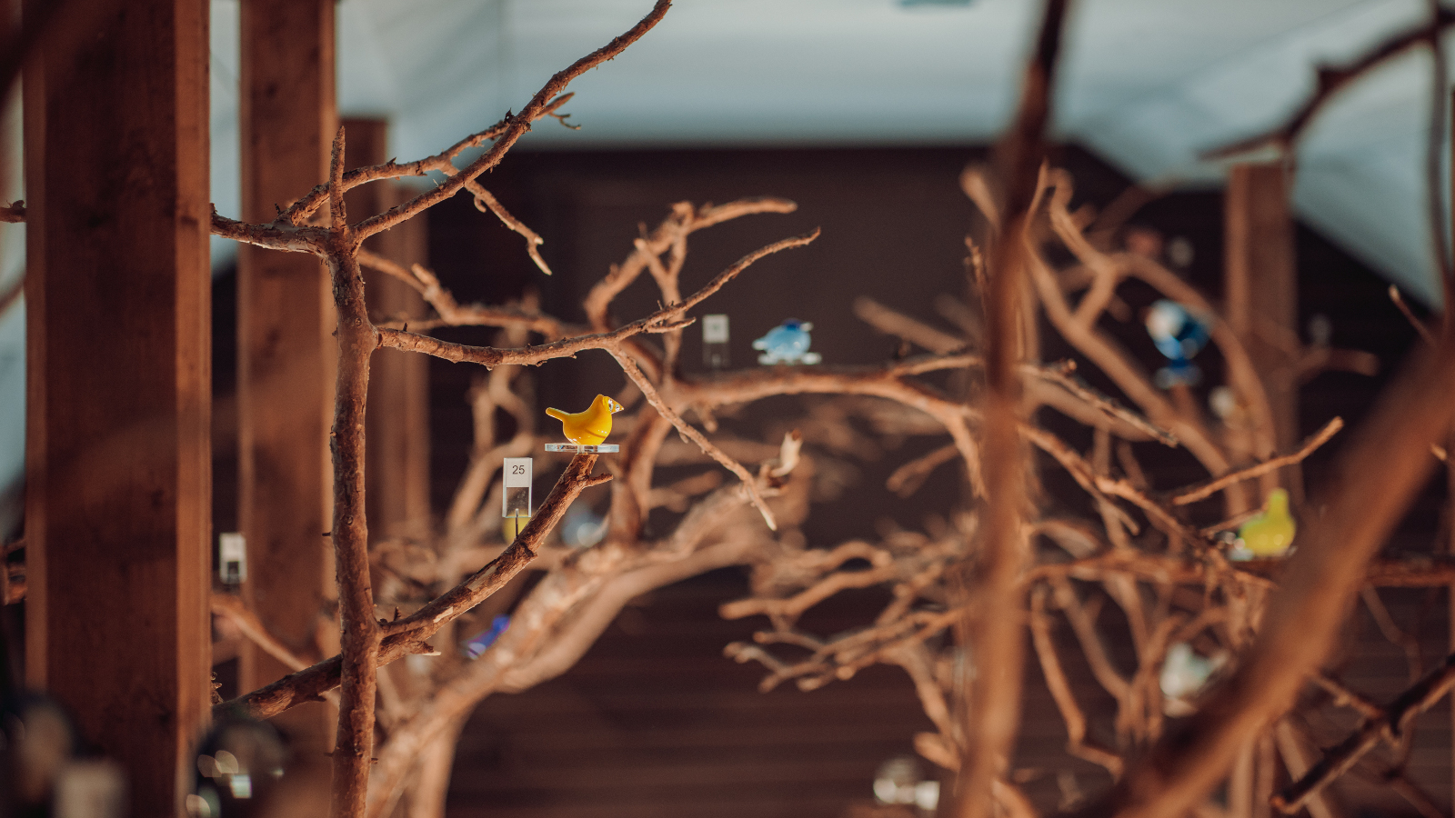 Kuvassa on ruskeissa pystyrungoissa ruskeita puun oksia, joissa on pieniä eri värisiä lasilintuja.