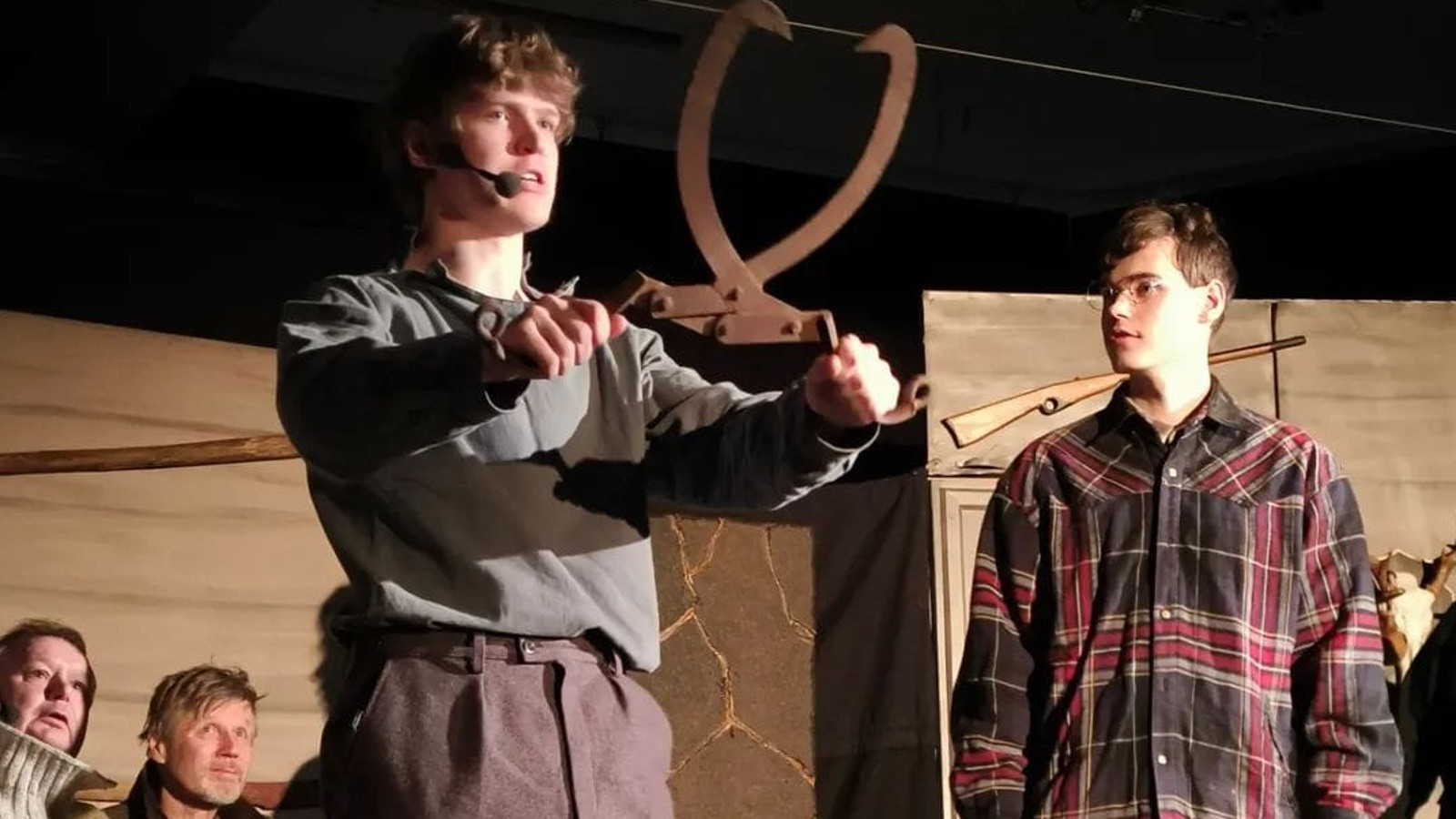 Kuvassa on vasemmalla nuori mies, joka pitää isoja hakasia käsissään ylöspäin. Vieressä oikealla on toinen nuori mies katsomassa ja taustalla istuu mies.