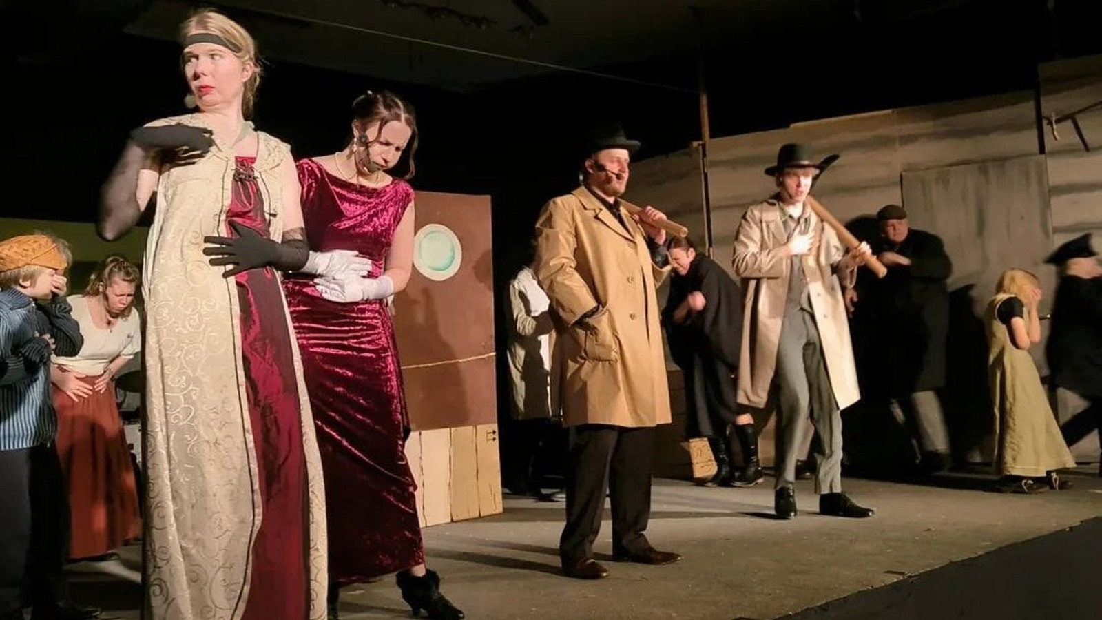 Kuvassa on näyttämöllä vasemmalla nuori nainen pitkässä puvussa, jossa on vaalea liivi ja kiiltävän punainen puku. Vieressä oikealla nuori nainen pitelee vatsaansa ja hänellä on punainen pitkä puku. Keskellä on kaksi miestä vaalean ruskeissa trensseissä laulamassa. Taustalla on runsaasti näyttelijöitä ja keskellä ruskea lavaste.