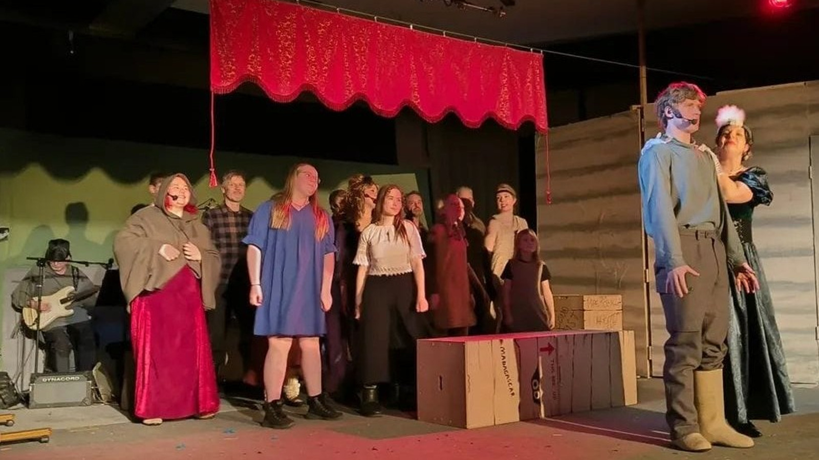 Kuvassa on oikealla näyttämön reunassa nuori mies ja nainen vihreässä pitkässä puvussa seisomassa vierekkäin ja nainen pitää miestä olkapäästä kiinni. Vasemmalta on takana iso joukko näyttelijöitä seisomassa ja laulamassa. Yläpuolella on punainen yläverho.