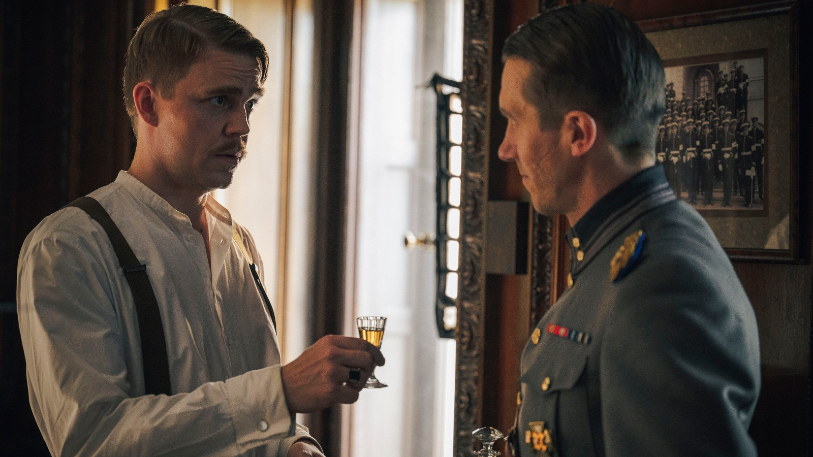 Kuvassa on vasemmalla Aku Sipola valkoisessa paidassa kohottamassa lasia sotilaspuvussa olevalla Jussi Vataselle, joka on osittain selin. Taustalla on iso ikkuna ja kuva sävy on tummahko. 