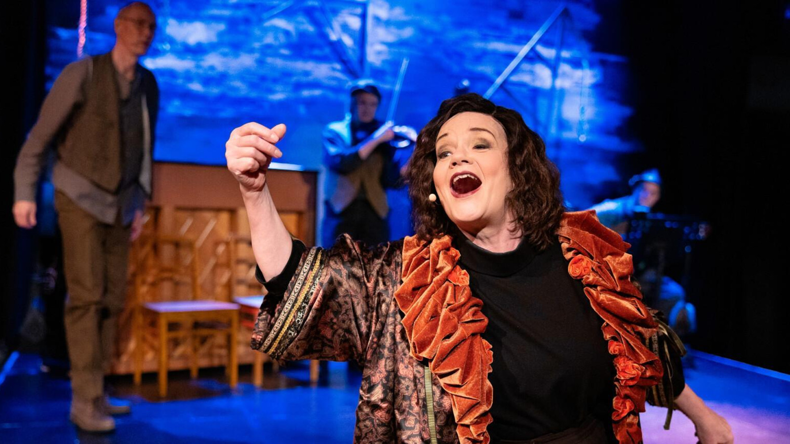 Kuvassa on Lise Holmberg keskellä laulamassa mustassa mekossä ja ruskehtavassa rimpsujakussa. Taustalla on vasemmalla mies ruskehtavissa vaatteissa ja takana on soittaja.