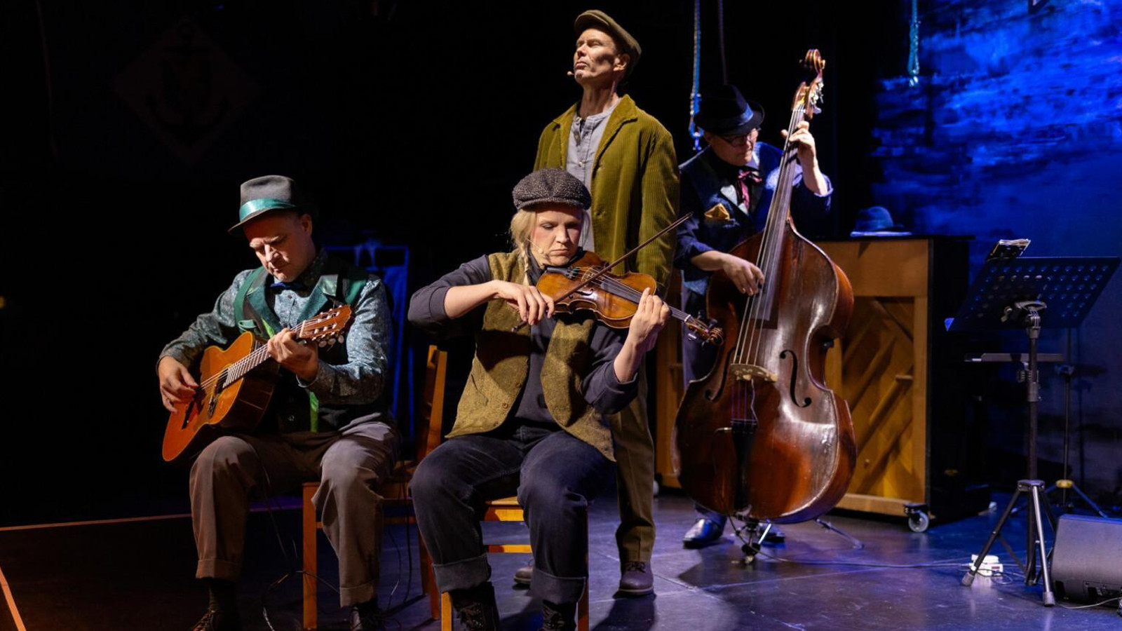 Kuvassa ovat soittajat ja vasemmalla on mies istumassa soittamassa kitaraa. Hänellä on sininen takki päällään ja hattu. Hänen vieressään oikealla nainen ruskeassa liivissä ja lippalakissa soittamassa viulua. Heidän takanaan on mies laulamassa seisoen ja häänellä on vihertävä pitkä takki päällään. Takana on mies soittamassa bassoa.