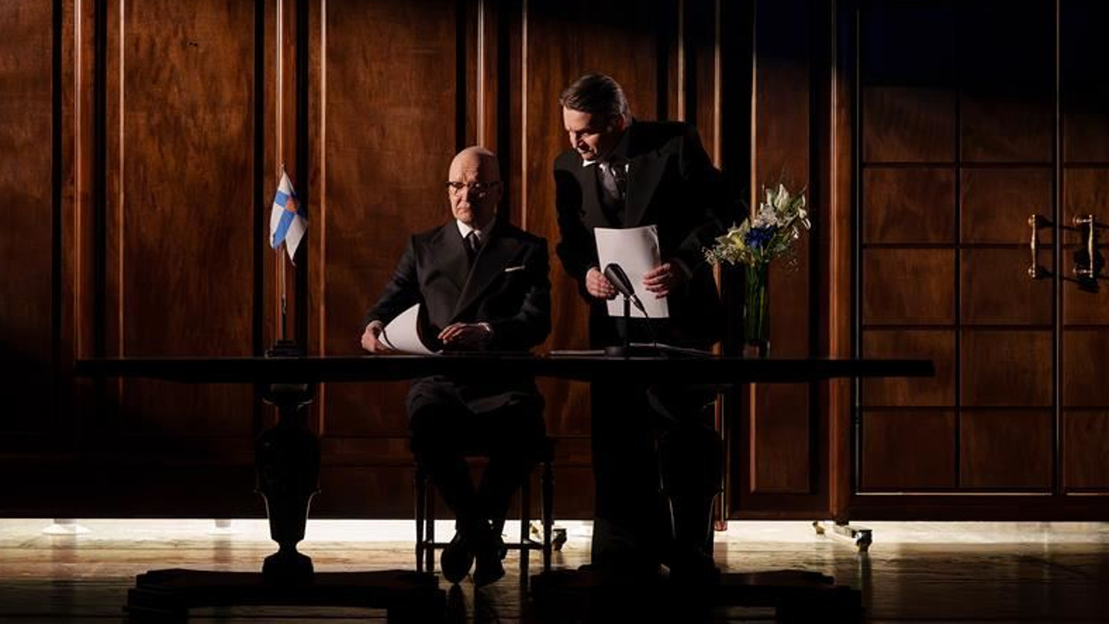 Kuvassa on tummasävyinen presidentin työhuone ja siinä tumma kirjoituspöytä, jossa on vasemmassa kulmassa Suomen lippu. Pöydän takana istuu Vesa Vierikko ja oikealla vieressä seisoo mies papereita kädessään.