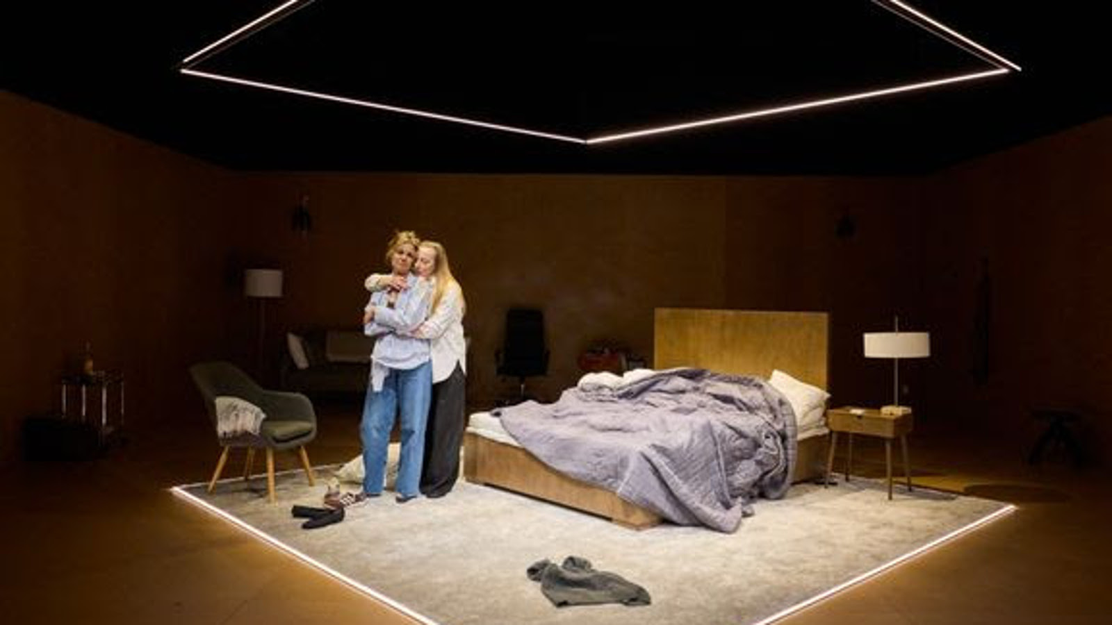 Kuvassa on valaistulla näyttämöllä, jossa on takana oleva pitkähiuksinen, valko-paitainen Milla Kangas halaa Anni-Maija Koskista, jolla on siniset farkut ja vaalean sininen paitapusero. Heidän takanaan on pari vuode, jossa on vaalean harmaa päiväpeitto sekaisin. 