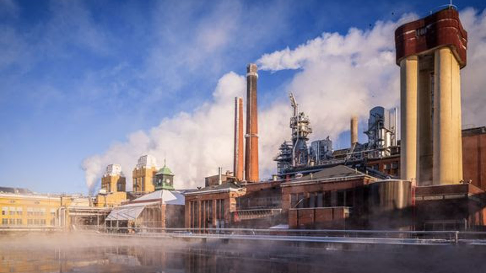 Kuvassa on edessä vettä ja takana punatiilisiä tehdasrakennuksia, joissa on torneja ja oikealla korkeat savupiiput, joista tulee savua. Tautalla on sinistä taivasta.