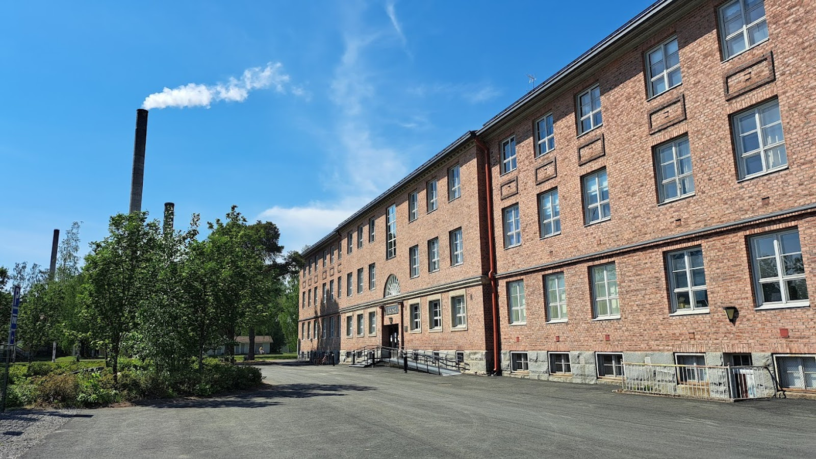 Kuvassa on oikealla punatiilinen kolmekerroksinen museorakennus.  Vasemmalla on lehtipuita ja kolme tehtaanpiippua. Taustalla on sinistä taivasta.