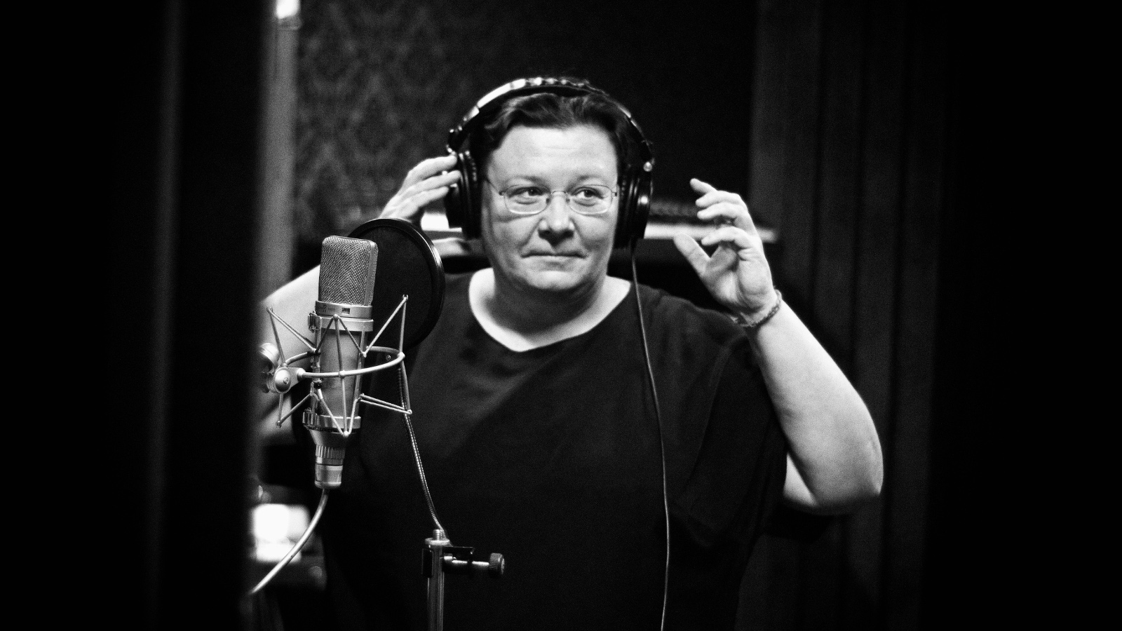  Kuvassa on Maija Kaunismaa kuulokkeet korvissaan äänitysstudiossa ja hän pitää käsiään niissä. Hänellä on musta pusero päällään. Kuva on musta-valkoinen.