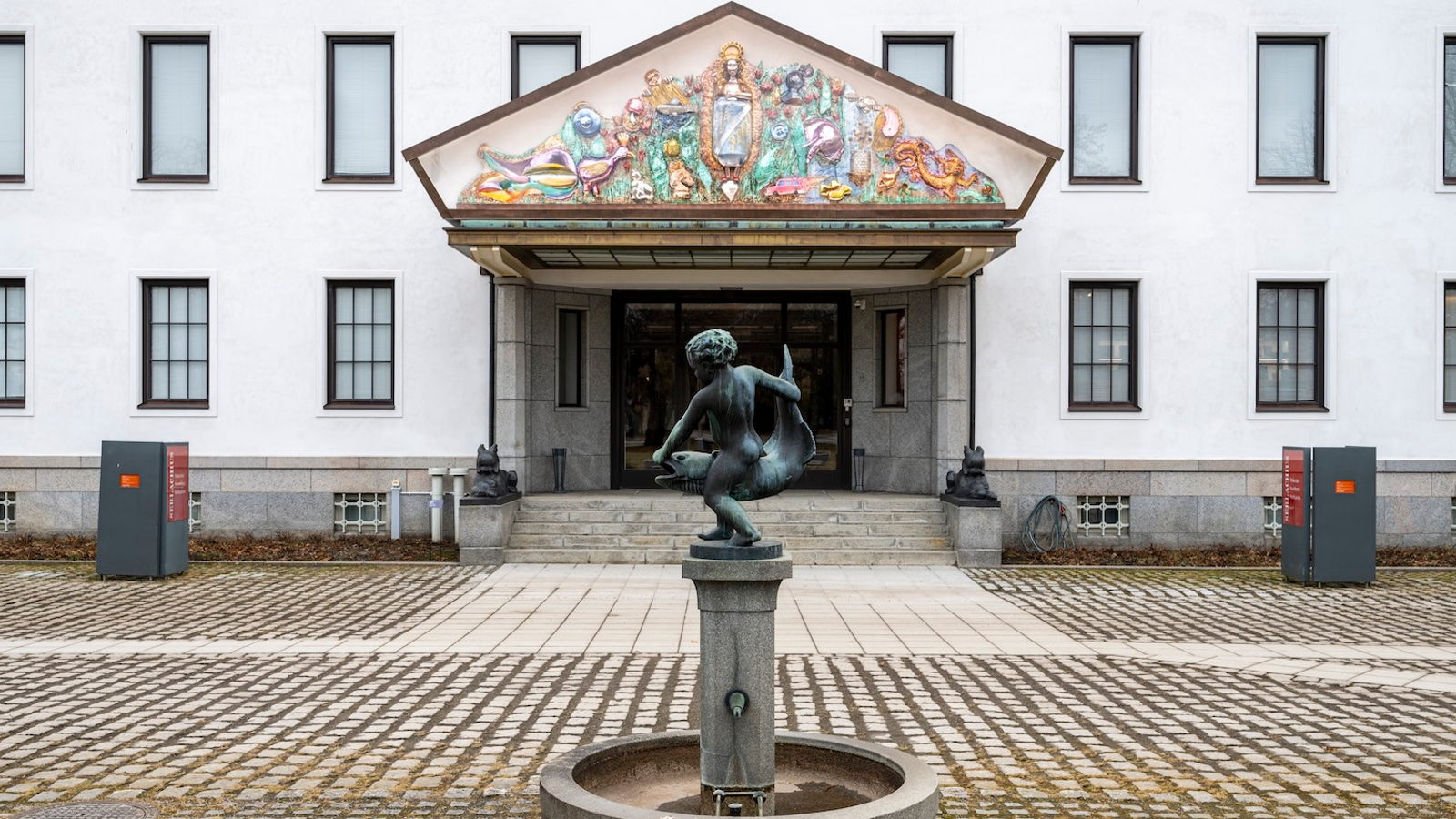 Kuvassa on valkoinen rakennus jossa on kapeita ikkunoita kahdessa kerroksessa. Siinä on portaikko ja isot ovet ja niiden päällä iso kolmio. Siinä on reliefimäisesti palstellivärisiä hahmoja ja esineitä.