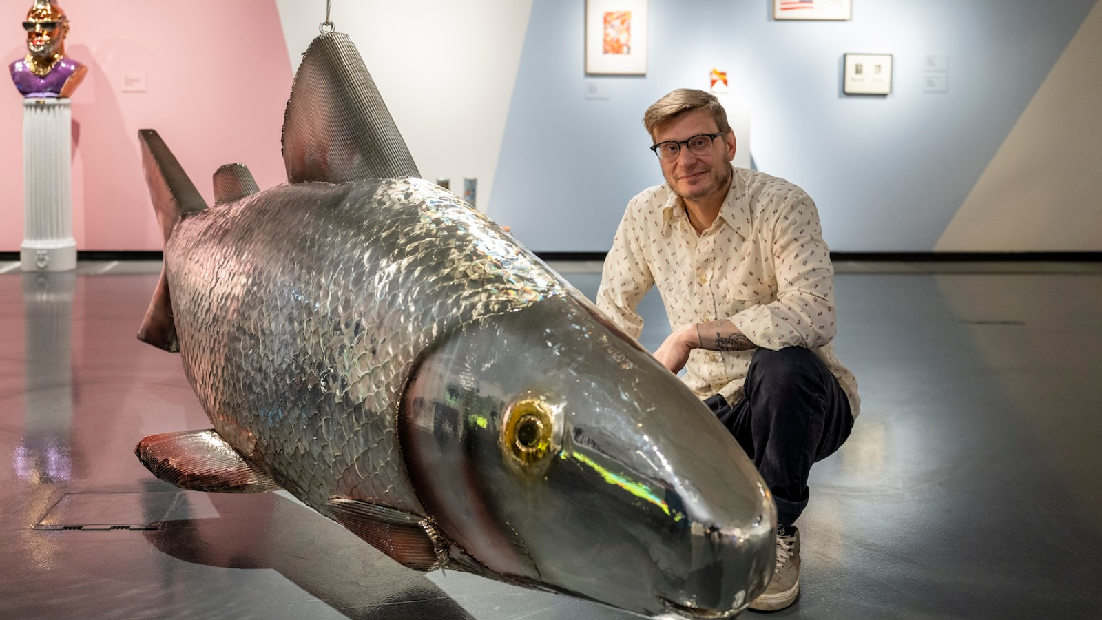 Kuvassa on näyttelytilassa hieman pikittain iso siika metallista. Vieressä on kyykyssä Anssi Kasitonni. Taustalla näkyy vasemmalla kuntainen muotokuva jalustan päällä ja seinällä tauluja.