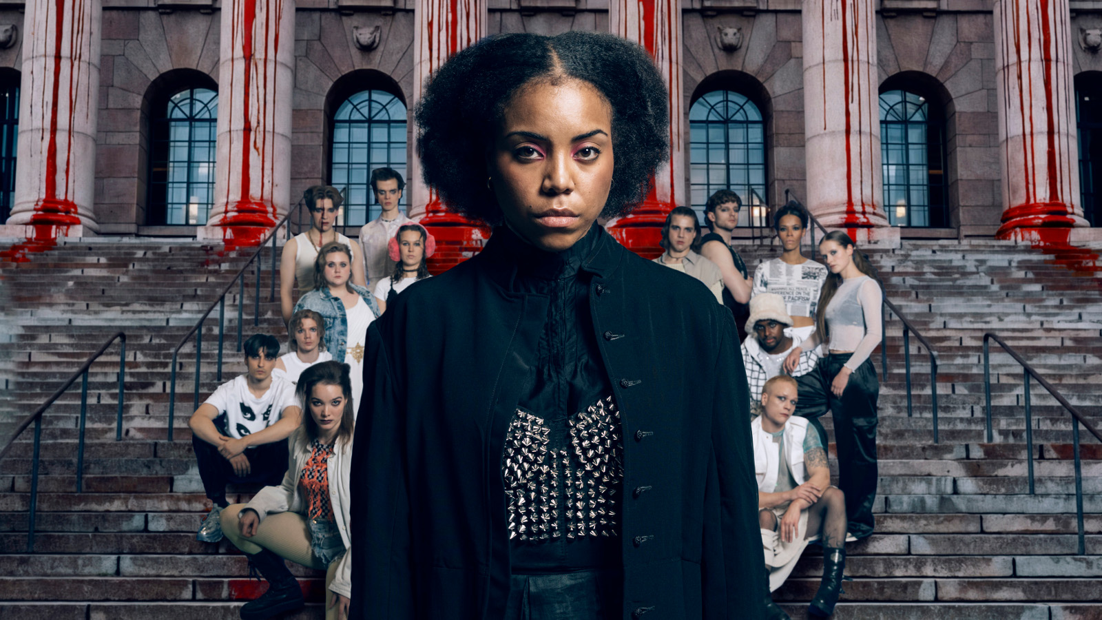 Kuvassa on keskellä nuori nainen mustassa kaavussa, jossa on hopeinen perhonen edessä. Hänellä on mustat, kiharat hiukset. Taustalla on valoisissa puseroissa istumassa harmailla portailla joukko nuoria. 