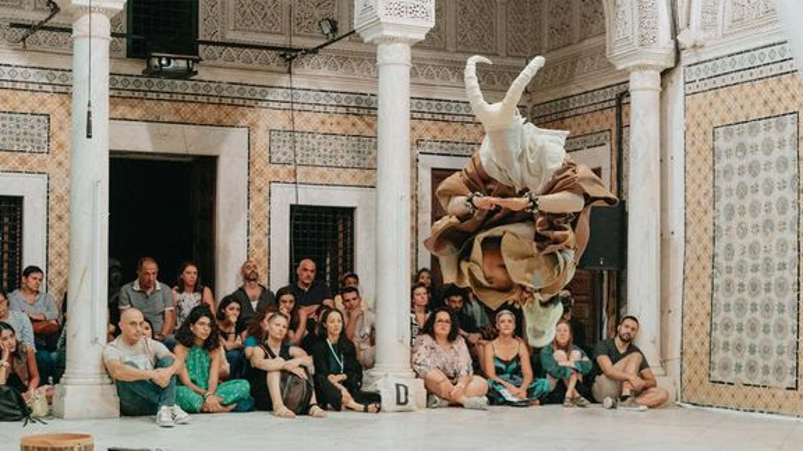 Kuvassa on joukko ihmisiä istumassa rakennuksen valkoisella lattialla ryhmänä ja edessä on kolme valkoista, itämaista pylvästä. taustalla näkyy seinää, jossa on valkoinen, avoin oviaukko ja vieressä kaksi pienempää aukkoa. Seinässä on keltakuvioista mosaiikkia ja sinikuvioista ornamenttia.