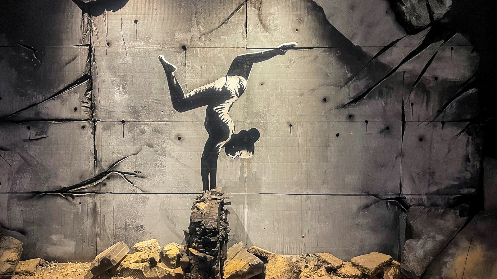 Kuvassa on ballerina seisomassa käsillään kivien päällä ja hänellä on jalat koukistettuna. Taustalla on harmaata betoniseinää. Maassa on vaalean ruskeita kiviä.