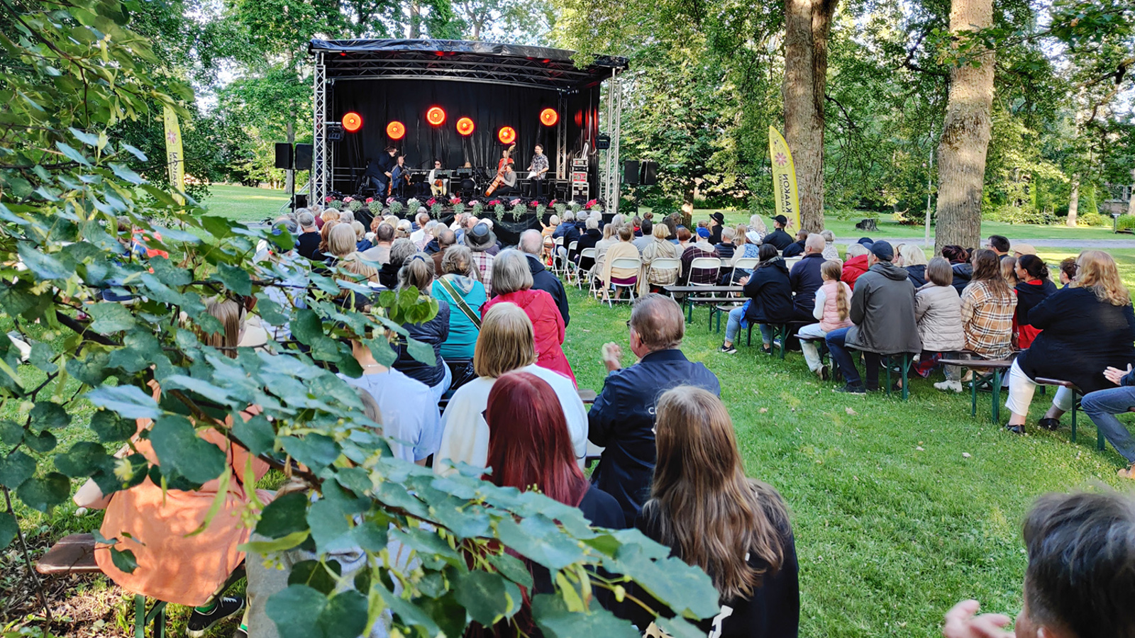 Kuvassa on puiston vihreitä puita ja oksia. Taustalla on esiintymislava ja siinä orkesteri soittamassa. Lavan edestä taakse päin on yleisöä istumassa tuoleilla.
