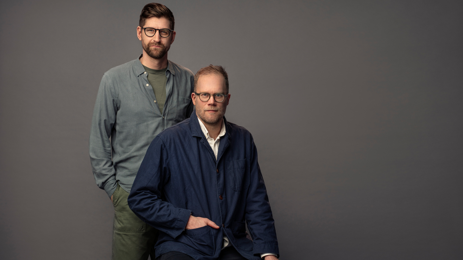 Kuvassa ovat Lars Berge seisomassa harmaassa puserossa Axel Gords Humlesjön takana ja Humlesjö istuu. Hänellä on musta pusero ja pitkät housut. Tausta on tumman harmaa.